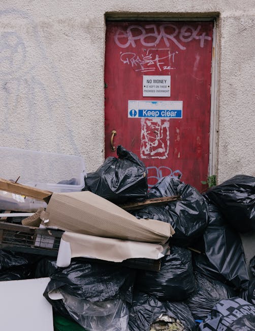 A collection of black garbage bags filled with household waste piled against a red metal door on an exterior wall, with some cardboard and discarded packaging scattered on top. The metal door features