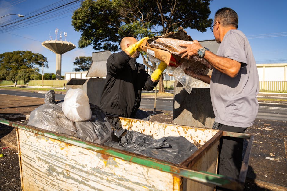 A person dressed in a grey outfit with a checkered yellow and grey jacket, wearing light green gloves, is holding open a large black rubbish bag made of plastic with a shiny finish. The bag appears to