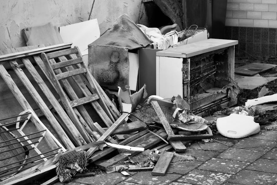 A narrow urban alleyway filled with accumulated waste and discarded rubbish, featuring a large black and white bag on a trolley in the foreground. Behind it, a substantial pile of cardboard boxes, pap